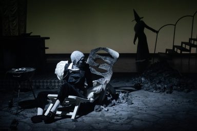Figur auf der Bühne, umgeben von Schatten und einer hintergründigen Hexen-Silhouette.