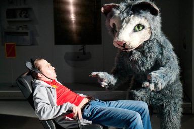 Ein Mensch sitzt in einem Stuhl, während eine menschengroße Katze ihn anstarrt.