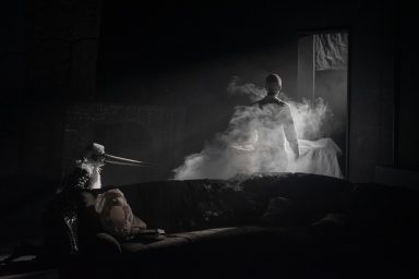 Eine von Nebel umhüllte Figur steht im Gegenlicht vor einer aufgebahrten Figur. Links daneben eine Figur mit langem Schnabel.