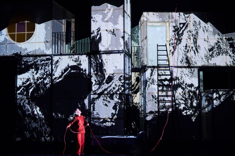 Frau Yamamoto ist noch da. Foto: Joseph Ruben Künstlerische Projektion des Marmolata Massivs auf ein Bühnenbild. Davor eine Schauspielerin in orangenem Anzug.
