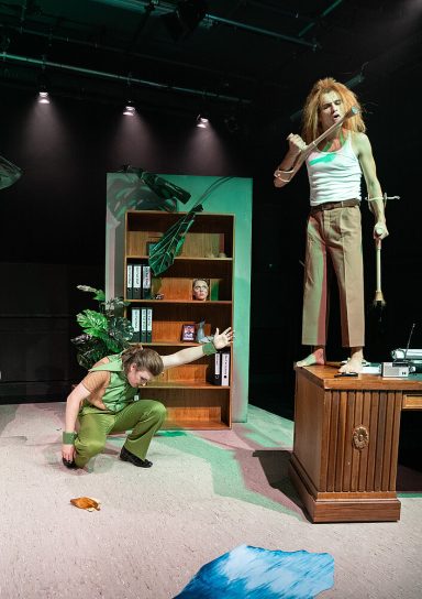 Zwei Schauspieler auf einer Theaterbühne, einer steht auf einem Schreibtisch, der andere kniet daneben.