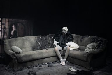 Mann in überzeichnetem Baby-Kostüm sitzt auf einer verbrannten Couch in einem dunklen Raum, trägt eine Mütze und sieht nachdenklich aus.