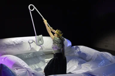 Eine Schauspielerin mit einer überdimensionalen Sicherheitsnadel auf einer nicht vollständig aufgeblasenen weißen Hüpfburg.