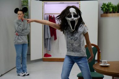 Person mit einer Maske und einem Schrei-Gesicht zeigt auf eine andere Person im Hintergrund.