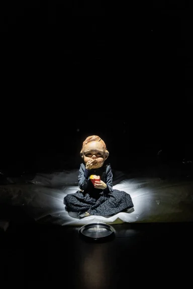 Schauspielerin mit Baby-Maske umgeben von dunklem Hintergrund und schimmerndem Licht isst Pommes.